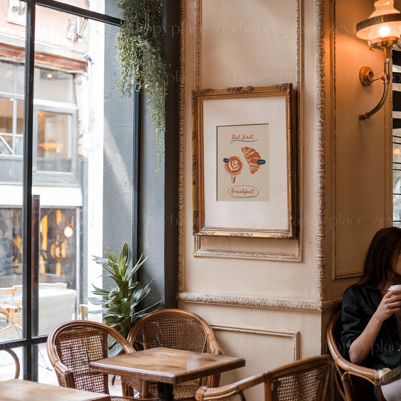 K&ouml;nnte beinhalten: Ein gerahmter Druck mit wei&szlig;em Hintergrund und einer bunten Illustration eines Croissants und einer Tasse Kaffee. Der Text auf dem Druck lautet "But first, breakfast". Der Druck h&auml;ngt an einer Wand in einem Caf&eacute;-Ambiente.