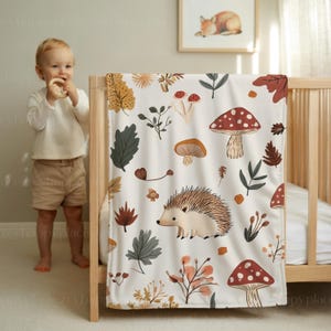 May include: A white baby blanket with a whimsical forest design. The blanket features illustrations of mushrooms, leaves, and a hedgehog in shades of brown, red, and green. The blanket is draped over a wooden cot.