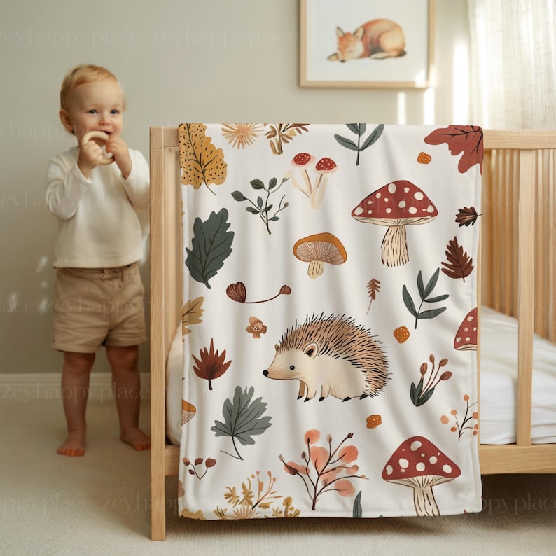 May include: A white baby blanket with a whimsical forest design. The blanket features illustrations of mushrooms, leaves, and a hedgehog in shades of brown, red, and green. The blanket is draped over a wooden cot.