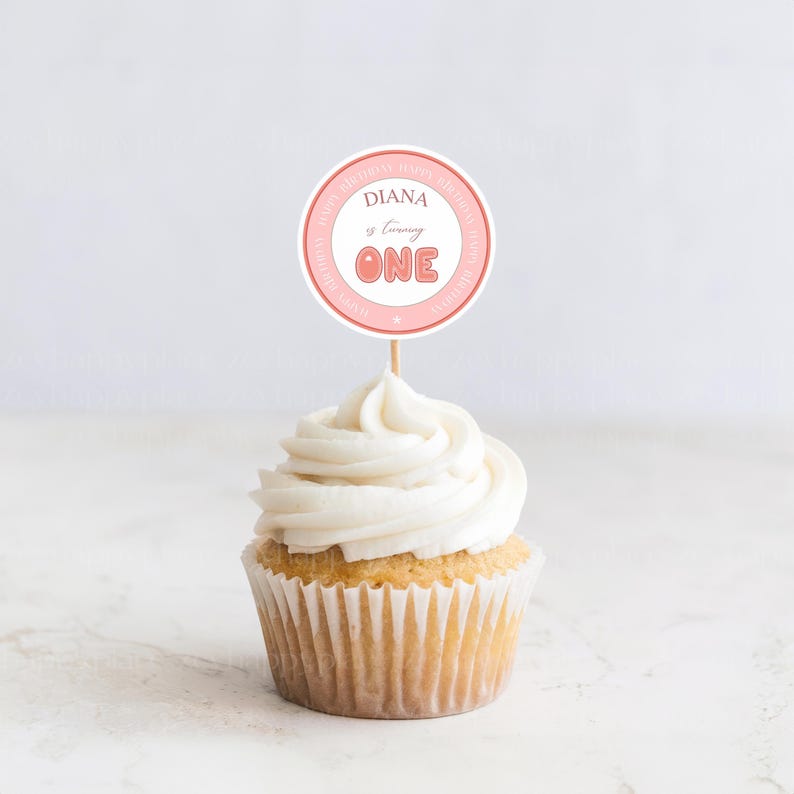 May include: Pink and white cupcake topper with the text "Diana is turning ONE" in a pink circle. The topper is on a cupcake with white frosting.