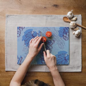 Op de afbeelding: Een persoon snijdt een rode tomaat op een blauw glazen snijplank met een druivenrankenontwerp. De snijplank ligt op een lichtgrijze linnen placemat.