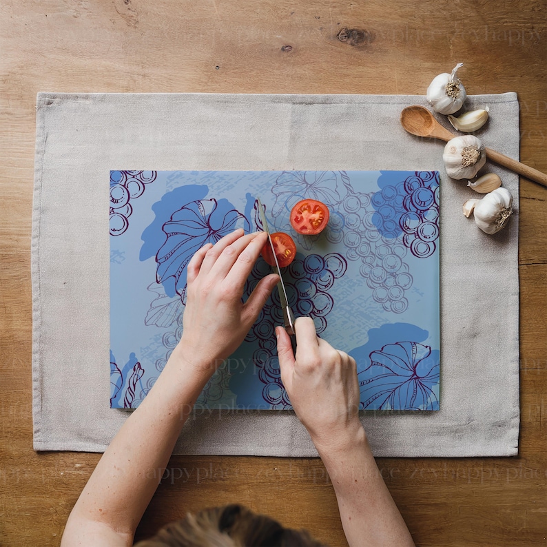 Op de afbeelding: Een persoon snijdt een rode tomaat op een blauw glazen snijplank met een druivenrankenontwerp. De snijplank ligt op een lichtgrijze linnen placemat.