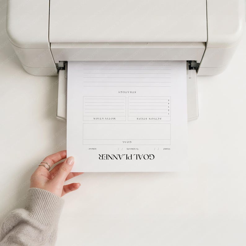 May include: A white printer printing a "GOAL PLANNER" sheet. The sheet has sections for strategy, motivation, and action steps. A hand holds the printed sheet.