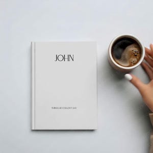 May include: A white journal with the name "JOHN" printed on the cover, along with the text "THINGS HE COULDN'T SAY." A brown mug filled with coffee sits next to the journal.