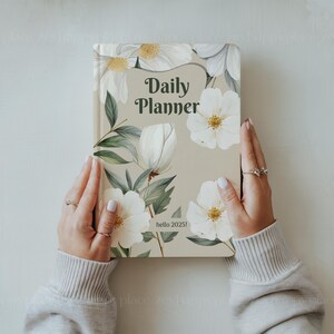 May include: A beige daily planner with a floral design featuring white flowers and green leaves. The text "Daily Planner" is printed in black on the front cover. The text "hello 2025!" is printed in black on the bottom of the cover.