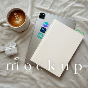 May include: A mockup scene featuring a white book, a silver tablet with app stickers, a speckled mug of latte art, and white wireless earbuds. The word "mockup" is displayed in a stylized font. The scene is set on a neutral-toned fabric.