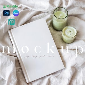 May include: A white book with the word "mockup" and file type details printed on the cover, resting on a white fabric surface. A green candle in a glass jar and a small cup of matcha are next to the book. The image includes the Printify, Ps, Canva, and ICC logos.
