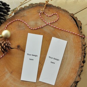 Puede incluir: Dos marcadores de p&aacute;gina blancos en blanco con el texto "Your design here" sobre un fondo de madera. Los marcadores de p&aacute;gina est&aacute;n atados con un cordel rojo y blanco.