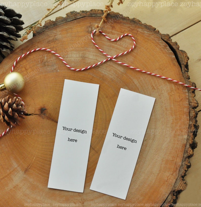 Puede incluir: Dos marcadores de p&aacute;gina blancos en blanco con el texto "Your design here" sobre un fondo de madera. Los marcadores de p&aacute;gina est&aacute;n atados con un cordel rojo y blanco.