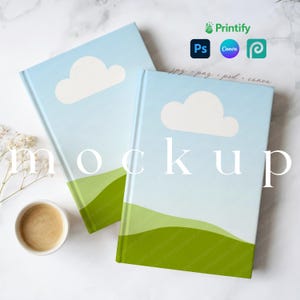 May include: Two hardcover notebooks with a light blue sky design, white cloud, and green hills. The word "mockup" is printed on the front. A cup of coffee and sprigs of white flowers are in the corner.