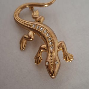 Könnte beinhalten: Eine goldfarbene Eidechsenbrosche mit geschwungenem Körper und langem Schwanz. Die Eidechse ist mit einer Reihe klarer, rund geschliffener Strasssteine entlang des Rückens sowie zwei kleineren Strasssteinen als Augen am Kopf verziert.