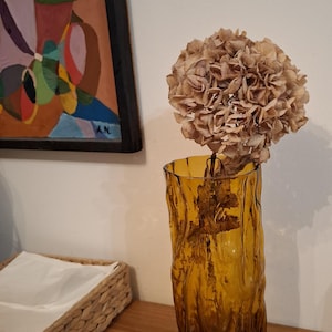 Könnte beinhalten: Eine hohe, zylindrische Vase aus bernsteinfarbenem Glas mit einer texturierten, gewellten Oberfläche. In der Vase befindet sich ein einzelner getrockneter Hortensienblütenkopf mit hellbraunen, papierartigen Blütenblättern.