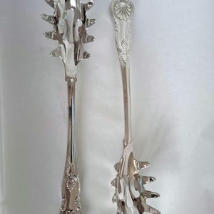 May include: A pair of silver-toned metal pasta serving utensils featuring a polished finish. Each tool has a long handle with an ornate shell-like motif at the base and a wide, pronged head designed for lifting noodles, showcasing a reflective surface.