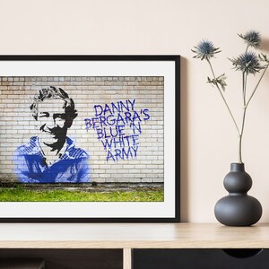 Puede incluir: Una impresión enmarcada de un mural de graffiti azul y blanco en una pared de ladrillo. El mural representa la cara de un hombre y el texto "Danny Bergara's Blue 'n White Army".