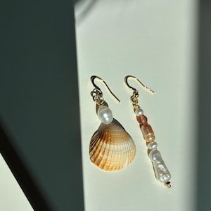 May include: A pair of mismatched earrings with gold-tone hooks. One earring features a single white freshwater pearl above a fan-shaped brown and white seashell. The other earring consists of a vertical strand of alternating white and peach pearls.