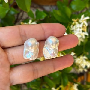 Peut inclure: Deux perles baroques blanches de forme irrégulière avec une finition irisée et très brillante. Chaque perle présente une forme organique unique avec des surfaces lisses et ondulées qui reflètent la lumière, révélant des textures nacrées.