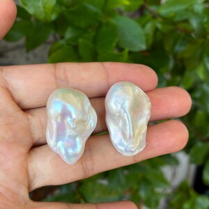 Peut inclure: Deux perles blanches de forme baroque et irrégulière avec une finition irisée très brillante. Chaque perle présente une texture de surface organique unique avec de subtils reflets de lumière rose et bleue sur leurs formes lisses et arrondies.