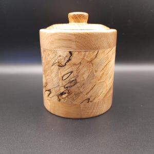 May include: A handcrafted wooden jar with a lid. The cylindrical jar has a light brown finish and a decorative band of lighter wood with dark markings. The lid has a small, rounded knob. The jar is likely used for storage.