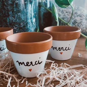 Peut inclure: Deux petits pots de fleurs en terre cuite avec une base peinte en blanc. Chaque pot porte l'inscription "merci" écrite en écriture cursive noire avec un petit cœur rouge en dessous. Les bords des pots sont naturels, couleur terre cuite.