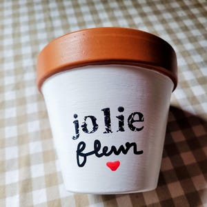 May include: A white terracotta plant pot with a wide, unpainted brown rim. The front features the black text "jolie fleur" in a distressed, stamped-style font, positioned above a small, solid red heart painted in a smooth, glossy finish.