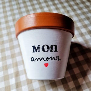 Puede incluir: Una maceta de terracota con el exterior pintado de blanco, que presenta el texto negro MON amour y un pequeño símbolo de corazón rojo. El borde de la maceta es de un color marrón rojizo natural y el cuerpo es cónico con acabado mate.