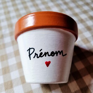 Op de afbeelding: Een kleine terracotta plantenpot met een wit geschilderd lichaam en een natuurlijke oranje rand. De voorkant van de pot bevat het woord Prénom in zwarte cursieve letters, geplaatst direct boven een klein, effen rood hartsymbool.