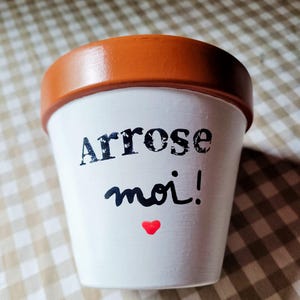 Peut inclure: Un pot de fleurs en terre cuite blanc avec un rebord marron. L'avant présente le texte noir Arrose moi! dans un mélange de polices serif et script, avec un petit symbole de cœur rouge positionné juste sous le texte sur la surface blanche.