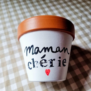 Pode incluir: Um vaso de plantas de terracota com o exterior pintado de branco, apresentando o texto em preto Maman chérie e um pequeno símbolo de coração vermelho. A borda do vaso mantém a sua cor natural de argila castanho-avermelhada, contrastando com o corpo branco.