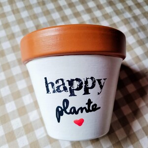 Può includere: Un vaso da fiori in terracotta con il corpo dipinto di bianco, caratterizzato dalla scritta nera "happy plante" e da un piccolo cuore rosso. Il bordo del vaso è di colore arancione terracotta naturale, mentre il corpo è bianco opaco.