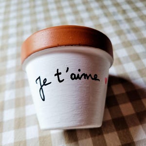 Pode incluir: Um pequeno vaso de plantas em terracota com o exterior pintado de branco, apresentando o texto escrito à mão em preto Je t'aime e um pequeno símbolo de coração vermelho na lateral. A borda do vaso mantém a cor natural da argila.