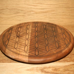 Peut inclure: Un dessous de verre rond en bois avec un motif de Fleur de Vie gravé en brun foncé. Le dessous de verre est en bois brun clair et présente une finition lisse et polie. Le motif est géométrique, fait de cercles qui se chevauchent.