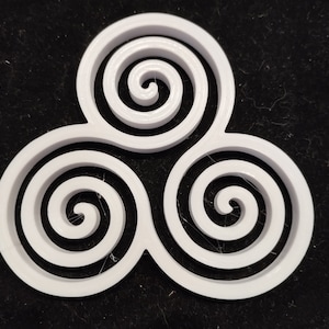May include: A white, three-part spiral design, possibly a decorative element or emblem, against a dark background. Each section features a concentric coil, creating a symmetrical and visually appealing pattern. The object is likely made of plastic.