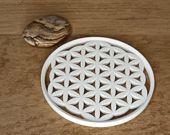 May include: White circular coaster with a raised flower of life design. The coaster is made of a smooth, white material and has a slightly raised edge.
