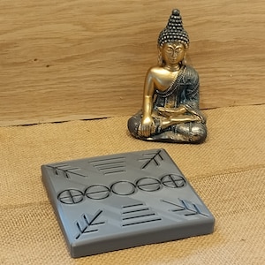 Peut inclure: Un carré gris avec un motif géométrique, comprenant des flèches et des cercles. Une statue dorée de Bouddha est posée sur une surface en toile de jute en arrière-plan.