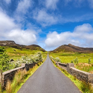 May include: A paved road winds through a green valley with rolling hills in the distance. The road is lined with stone walls and trees. The sky is blue with white clouds.
