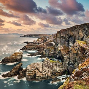 Puede incluir: Un paisaje marítimo dramático con acantilados rocosos y un mar tormentoso. El cielo es una mezcla de nubes rosadas, moradas y grises. Los acantilados son irregulares y erosionados, y el mar está agitado y con olas blancas.