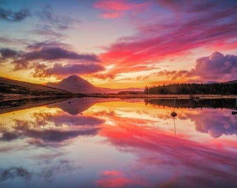 May include: A vibrant sunset over a still lake, reflecting the colorful sky in its surface. A mountain peak rises in the distance, creating a picturesque landscape.
