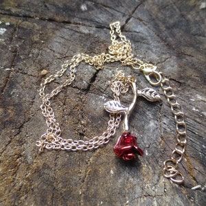 Puede incluir: Un collar dorado con un colgante de rosa. La rosa tiene una flor roja y hojas plateadas. El collar se muestra sobre una superficie de madera rústica y envejecida.