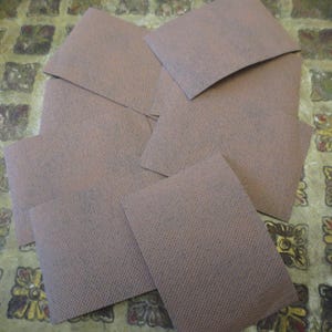 Può includere: Un set di otto quadrati di tessuto marrone, forse per l'artigianato o il quilting. I quadrati sono disposti in uno schema casuale su una superficie con un motivo dorato e marrone.