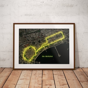 Op de afbeelding: Ingelijste print van het Baku City Circuit, een Formule 1-racecircuit in Azerbeidzjan. De lay-out van de baan is gemarkeerd in neongeel tegen een gedetailleerd luchtfoto van de stad. De print zit in een bruine lijst.
