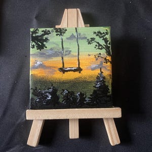Puede incluir: Una pintura en miniatura sobre un pequeño caballete de madera. La obra representa un paisaje sereno con un columpio colgando entre dos árboles, sobre un cielo de puesta de sol con tonos amarillos, naranjas y verdes. La pintura está enmarcada por siluetas negras de árboles.