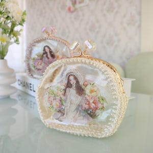 Puede incluir: Dos monederos color crema con cierres dorados y detalles decorativos de cristal. Cada monedero presenta una imagen impresa de una mujer rodeada de flores. Los monederos tienen un ribete de lentejuelas y se exhiben sobre una superficie reflectante.