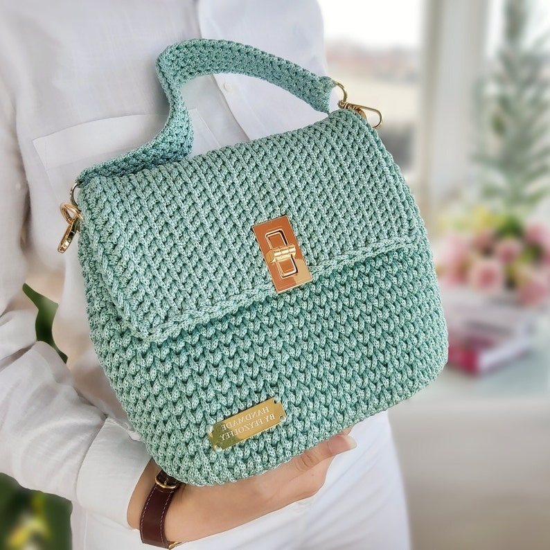 May include: A light green crocheted handbag with a gold clasp and a gold tag that reads "HANDMADE BY RENOVIERA".