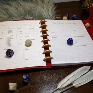 Puede incluir: Un cuaderno rojo con anillas, con páginas blancas y un patrón de cuadrícula. El cuaderno está abierto en una página con una hoja de personaje para un juego de rol. Hay varios dados azules y blancos esparcidos alrededor del cuaderno.