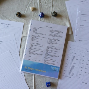 May include: An A5 Player Notebook with a blue and white design, surrounded by loose-leaf pages with tracker templates and dice. The notebook features a cheat sheet and the text "aurora mancy". Ideal for tabletop role-playing games.