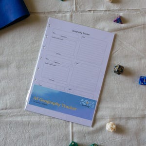 Könnte beinhalten: Ein weißes A5 Geography Tracker Papierblatt mit schwarz gedruckten Feldern für Notizen, Organisationen und Koordinaten. Unten befindet sich ein blaues Banner mit dem Text A5 Geography Tracker und aurorsmancy gaming. Mehrere mehrseitige Würfel liegen verstreut.