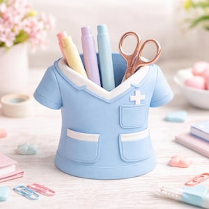 Peut inclure: Un organiseur de bureau bleu clair en forme de blouse m&eacute;dicale, contenant des stylos et des ciseaux dor&eacute;s. L'organisateur a des d&eacute;tails blancs et un embl&egrave;me de croix. Autres accessoires de bureau : un r&eacute;veil rose et des cahiers.