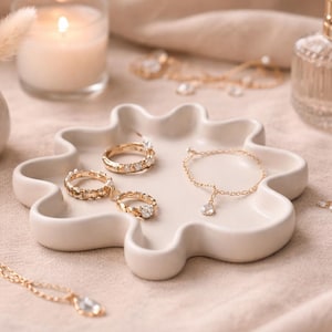 Op de afbeelding: Een verzameling gouden sieraden, waaronder ringen, een armband en een ketting, gepresenteerd op een crème-kleurig, bloemvormig dienblad. Een brandende kaars, een parfumflesje en een kleine vaas met gedroogde bloemen zijn ook te zien. De sfeer is zacht en elegant.