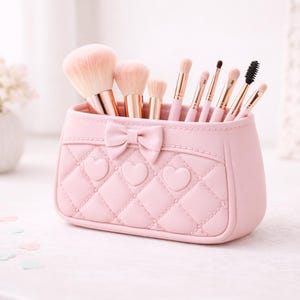Op de afbeelding: Een roze make-up kwastenhouder in de vorm van een tas, met strik- en hartdetails. De houder bevat verschillende make-up kwasten met roze handvatten en gouden hulzen. Op de achtergrond een vaas met bloemen en een decoratieve spiegel.