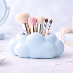 K&ouml;nnte beinhalten: Ein hellblaues, wolkenf&ouml;rmiges Make-up-Pinsel-Halter mit verschiedenen Make-up-Pinseln. Die Pinsel haben hellfarbene Borsten und metallische Griffe. Der Hintergrund umfasst einen Spiegel, Parf&uuml;m und Perlen, was eine weiche, pastellfarbene &Auml;sthetik erzeugt.